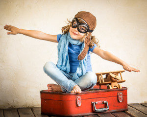 little girl sitting on old red suitcase pretending to fly, wearing pilot goggles and sitting next to wooden toy plane